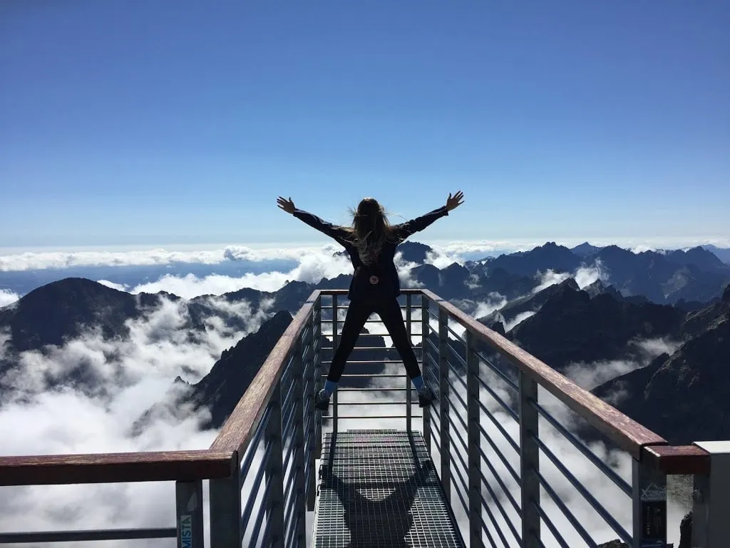 Person standing with arms raised on a mountain overlook above clouds and peaks